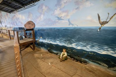 Susan Grove sits next to her mural at the St. George Dinosaur Discovery Site, St. George, Utah, date unspecified | Photo courtesy of Susan Grove, St. George News